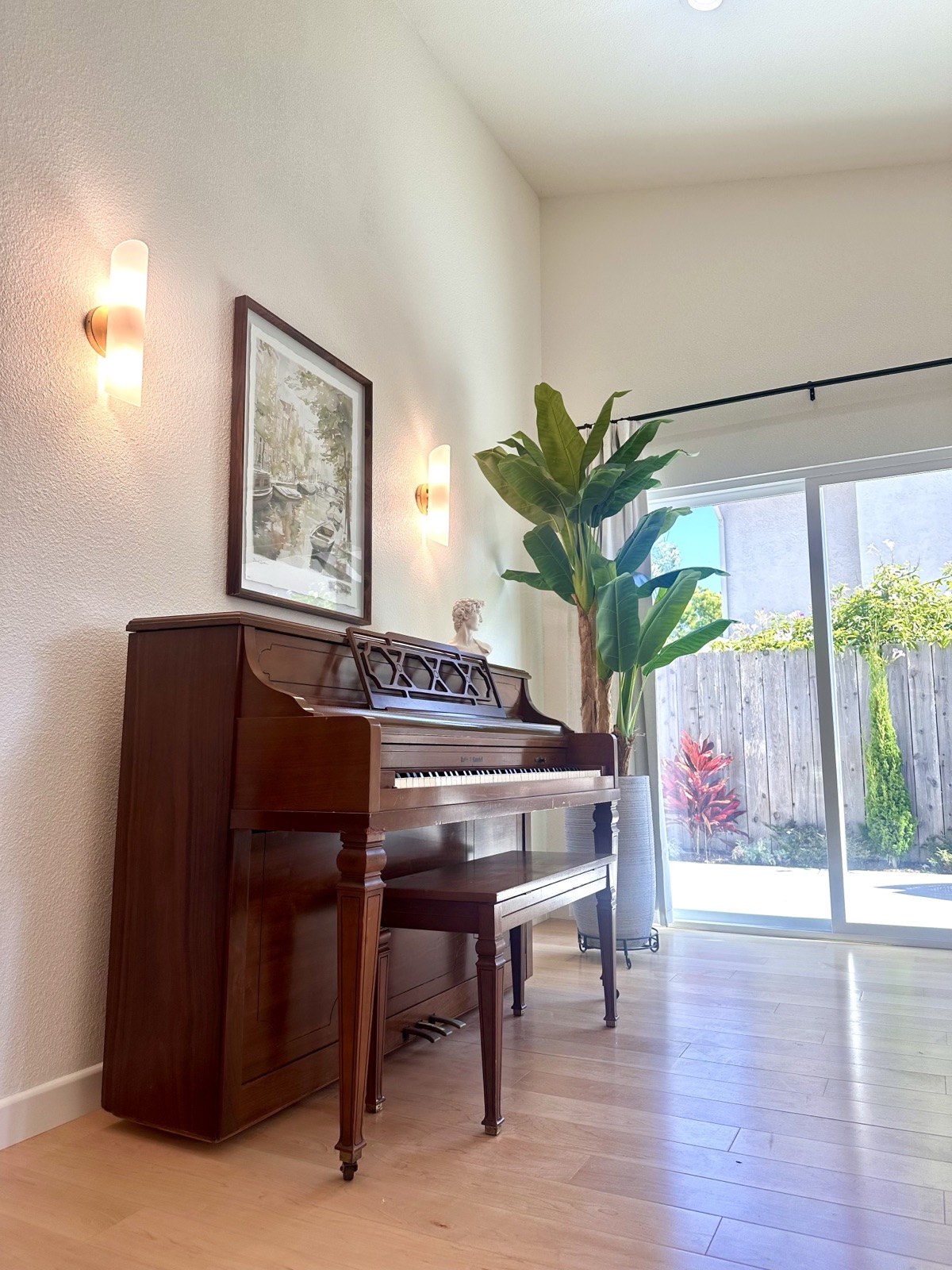 Upright piano beneath lit wall sconces beside a garden-view sliding door
